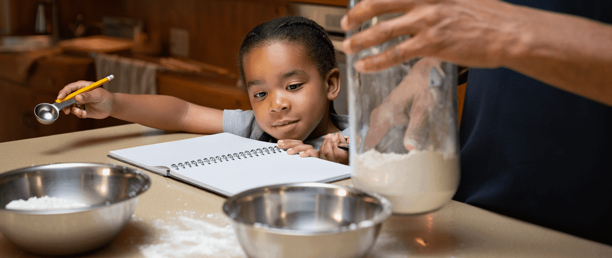 Child holding a measuring spoon, adult's hand in flour jar, bowls, and notebook.