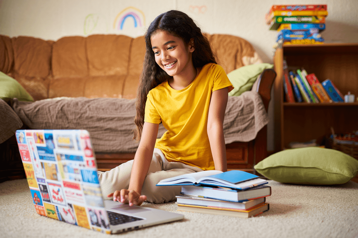 Smiling child uses a laptop on the floor with books nearby.