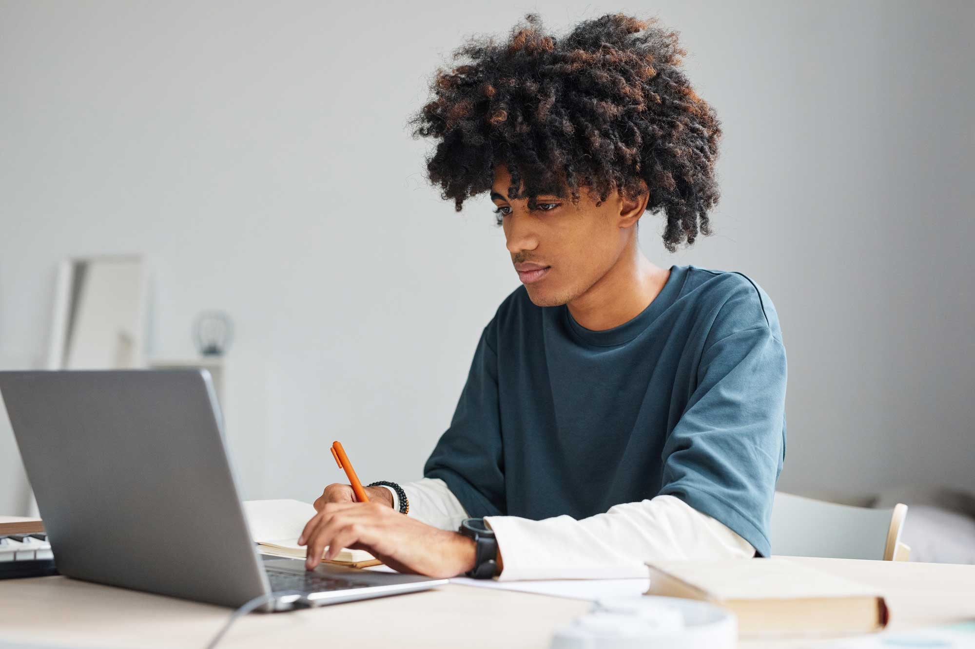 Young adult using laptop and writing in a notebook.