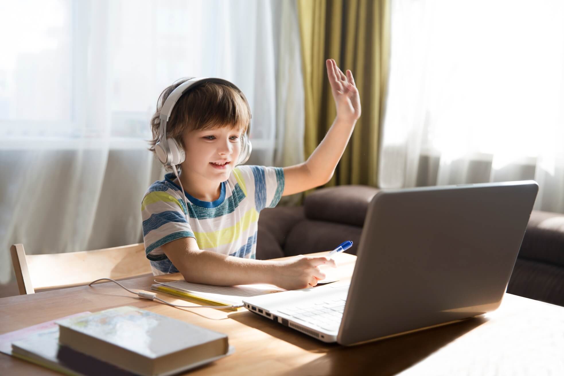 A home-schooled child sitting at a desk with a laptop, hand raised